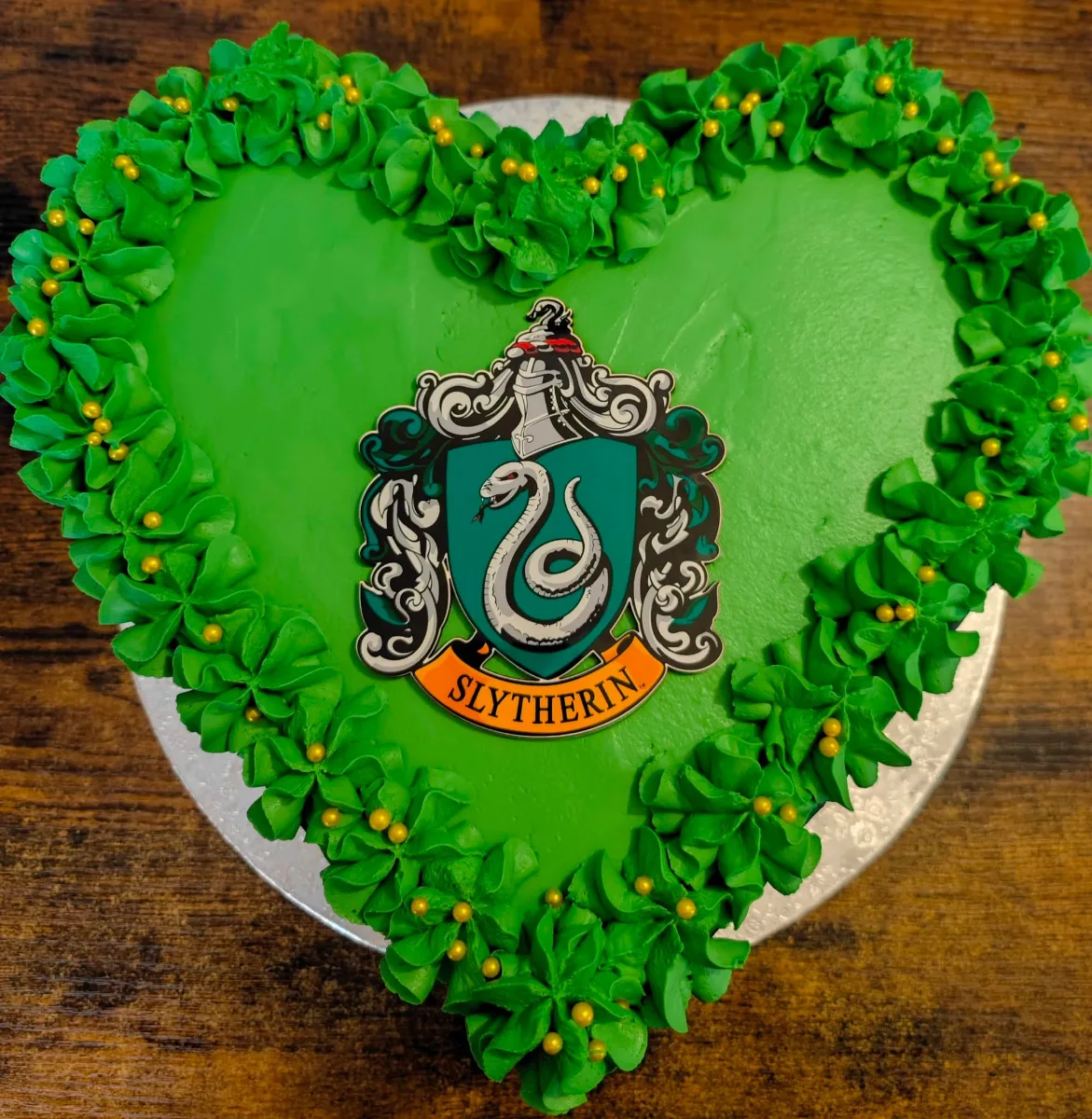 Srdcový zelený dort se zápichem s erbem Slytherinu uprostřed, pipovaný okraj a zlaté drobné ozdoby.